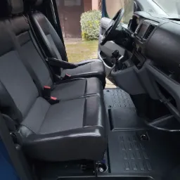 Wnętrze kabiny busa z ciemną tapicerką, widok na fotele kierowcy i pasażera, czarna podłoga z wytłoczonym wzorem.