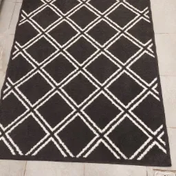 Ciemnoszary dywan z geometrycznym wzorem romb&oacute;w w kolorze białym, leżący na płytkach podłogowych w odcieniach beżu i szarości, widoczny z g&oacute;ry, z kratką odpływową w prawym dolnym rogu.