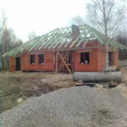 Surowy stan budowy domu z czerwonej cegły w Węgierskiej Górce, z widoczną konstrukcją dachu z zielonych belek i stertą żwiru na pierwszym planie. Widoczne drewniane podpory. Budynek w stanie...