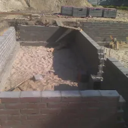 Budowa fundamentów domu murowanego z betonowych bloczków, widoczny wykop wypełniony piaskiem i fragmenty murów w różnym stadium zaawansowania.