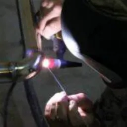 Praca spawacza: zbliżenie na proces spawania rury metalowej metodą TIG, widoczna jarząca się spoina i elektroda, w tle ciemne otoczenie warsztatowe.