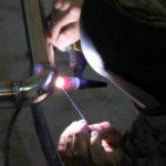 Praca spawacza: zbliżenie na proces spawania rury metalowej metodą TIG, widoczna jarząca się spoina i elektroda, w tle ciemne otoczenie warsztatowe.