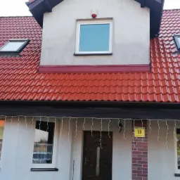 Dom jednorodzinny z nowym, błyszczącym, czerwonym dachem z dwoma oknami dachowymi i oknem na poddaszu, ozdobiony świątecznymi lampkami zwisającymi z rynny.