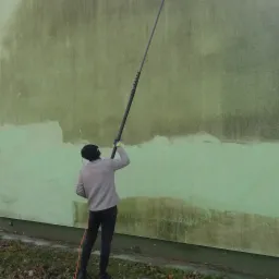 Pracownik w czapce i szarej bluzie czyści zieloną elewację budynku za pomocą lancy ciśnieniowej, widoczny efekt czyszczenia oraz wózek z urządzeniem czyszczącym na trawniku.