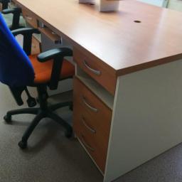 Pk Stolarstwo Meblowe - Długi, jasny stół z szufladami i otworem na kable, otoczony krzesłami biurowymi w pomieszczeniu z wyposażeniem laboratoryjnym w tle.