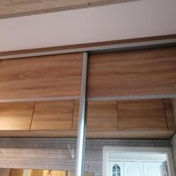 Pk Stolarstwo Meblowe - Zamknięta szafa wnękowa z przesuwnymi drzwiami z płyty drewnopodobnej, z widocznymi półkami wewnątrz i odbiciem wnętrza pokoju w lustrzanych drzwiach.