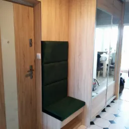 Wnęka z siedziskiem i tapicerowanym oparciem w kolorze butelkowej zieleni, obok drzwi wejściowych i szafy przesuwnej z lustrem, wykończona w okleinie drewnopodobnej.