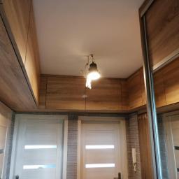 Pk Stolarstwo Meblowe - Wnętrze przedpokoju z zabudową szafek pod sufitem w kolorze drewna, dwoma białymi drzwiami z przeszkleniami i przesuwną szafą z lustrem.