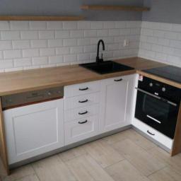 Pk Stolarstwo Meblowe - Nowoczesna kuchnia w narożniku: białe szafki z frezowanymi frontami, drewniany blat, czarny zlew i bateria, płytki typu metro na ścianie, drewnopodobna podłoga, piekarnik w zabudowie.