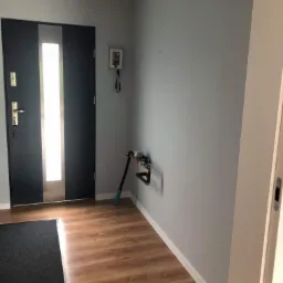 Nowoczesna, grafitowa dwuskrzydłowa drzwi wejściowe z przeszkleniem, widok na przedpokój z panelami podłogowymi, szarą ścianą i wystającym licznikiem wody.