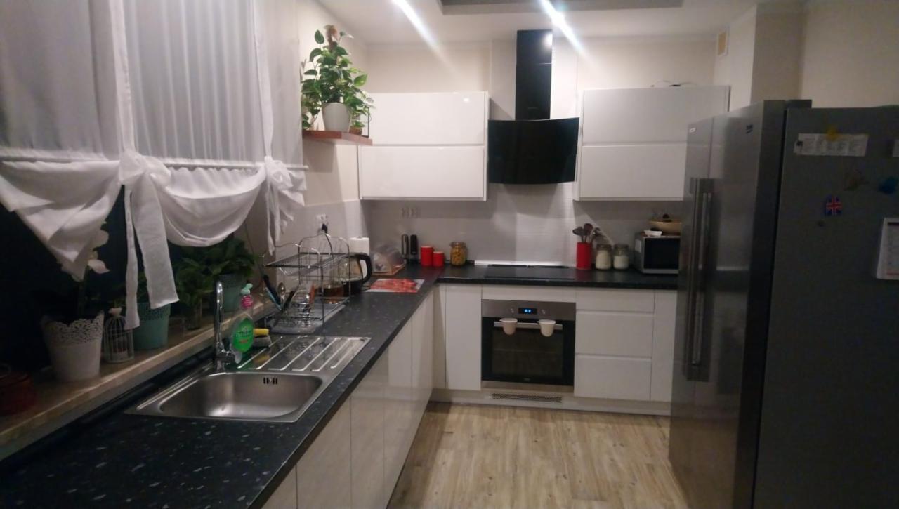 Jasna kuchnia z białymi szafkami i czarnym blatem w Jastrzębiu-Zdroju, widok na okno z firankami i stalowy zlew.