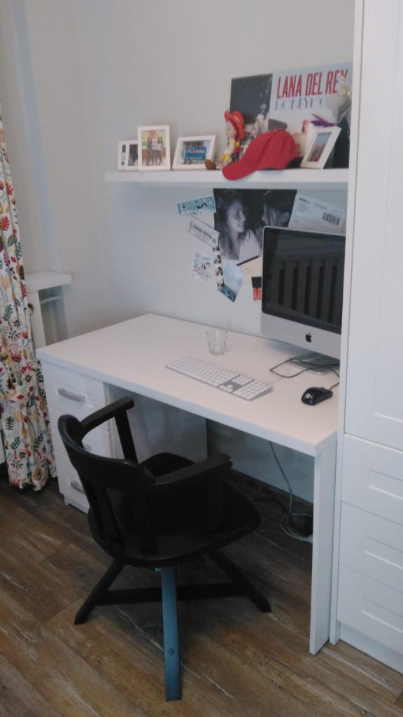 Białe biurko z szufladami i komputerem Apple, czarne krzesło na drewnianej podłodze, półka nad biurkiem z dekoracjami i zdjęciami, obok biała szafa.