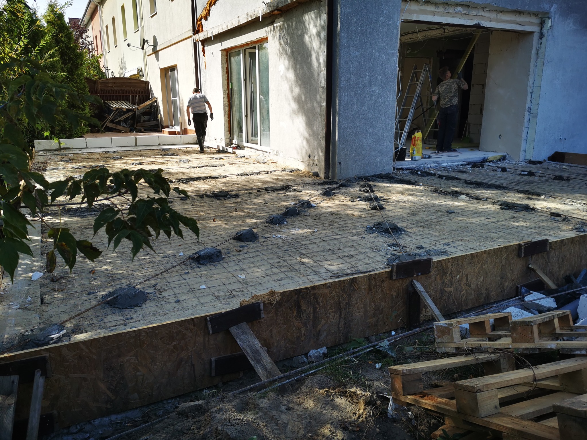 Przygotowanie podłoża pod wylewkę na zewnątrz budynku, widoczna siatka zbrojeniowa, szalunki z desek, palety oraz pracownik w tle.