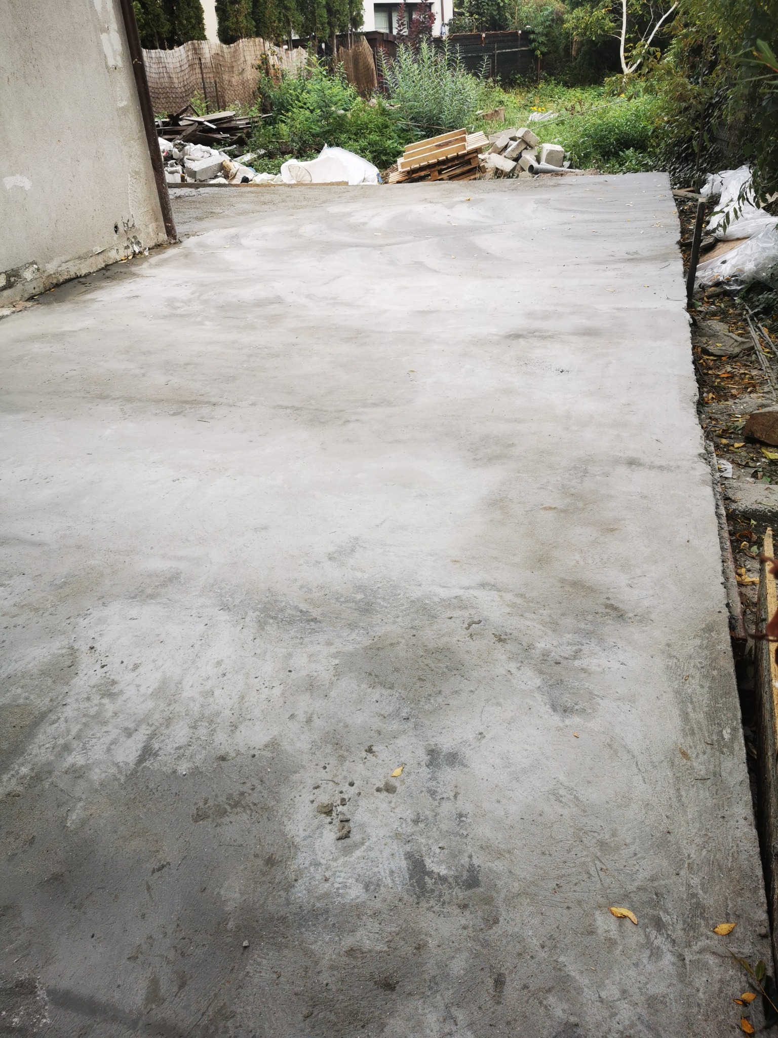 Świeżo wylana, szara betonowa wylewka na zewnątrz, z widocznymi śladami po narzędziach, otoczona zielenią i materiałami budowlanymi.