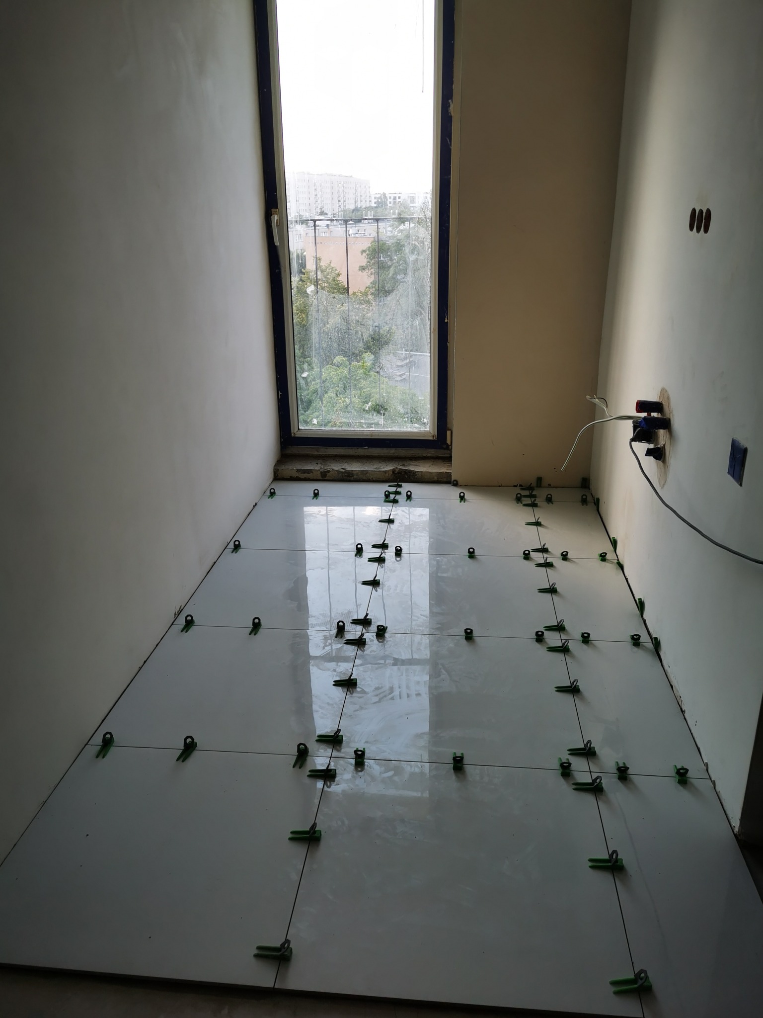 Układanie białych płytek podłogowych z użyciem zielonych krzyżyków poziomujących, widok na okno z budynkami w tle.