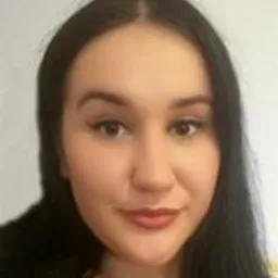 Portret kobiety o ciemnych włosach i delikatnym makijażu, uśmiechająca się lekko w kierunku kamery, na jasnym, rozmytym tle.