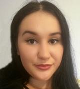 Portret kobiety o ciemnych włosach i delikatnym makijażu, uśmiechająca się lekko w kierunku kamery, na jasnym, rozmytym tle.