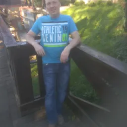 Mężczyzna w niebieskiej koszulce opiera się o drewnianą barierkę na tle zieleni i fragmentu starego młyna.