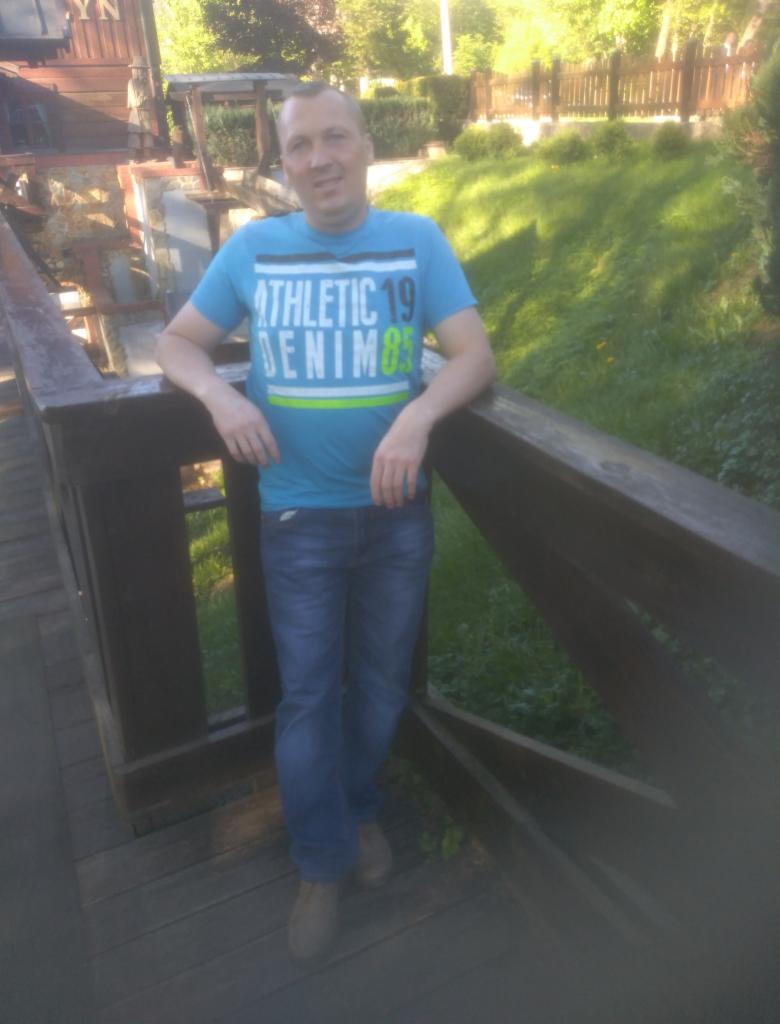 Mężczyzna w niebieskiej koszulce opiera się o drewnianą barierkę na tle zieleni i fragmentu starego młyna.