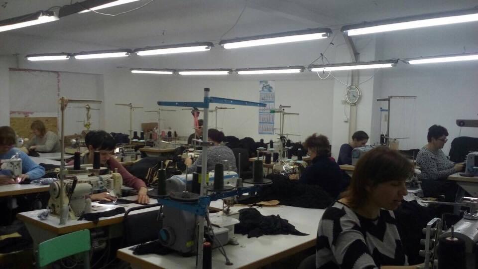 Wnętrze hali produkcyjnej z rzędem stanowisk pracy z maszynami do szycia, przy których pracują kobiety. W tle widoczne regały z nićmi i kalendarz.