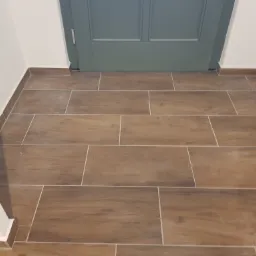 Ułożona podłoga z płytek imitujących drewno w przedpokoju, widoczna szara ościeżnica drzwiowa i białe ściany.