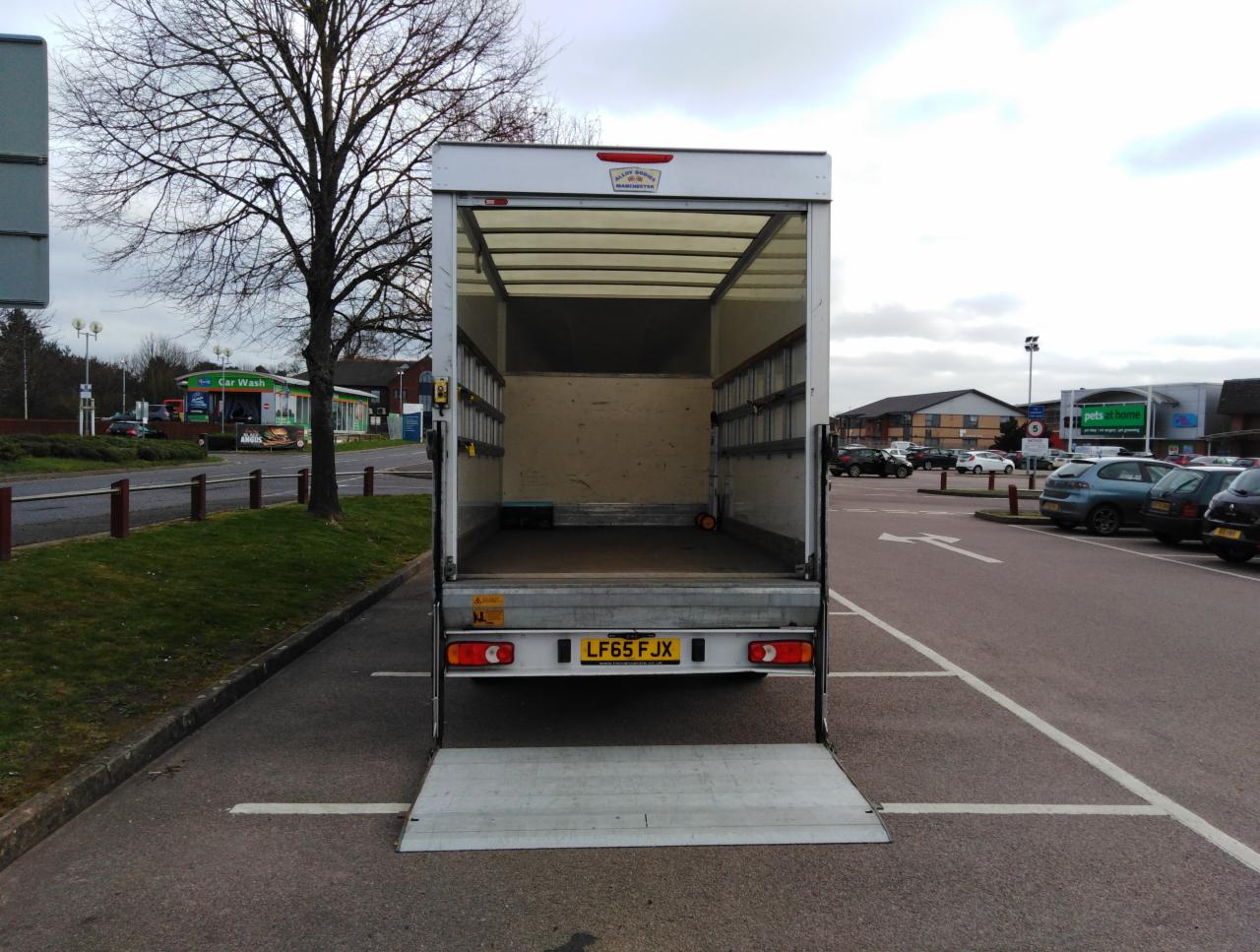 Pusta przestrzeń ładunkowa samochodu dostawczego z otwartą rampą, zaparkowanego na parkingu przed sklepami.