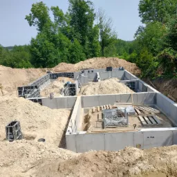 Wylany fundament domu jednorodzinnego z widocznymi elementami szalunkowymi i zbrojeniem, otoczony wykopem i ziemią, w tle zieleń drzew.