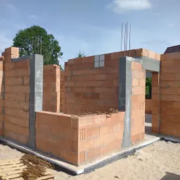 Wznoszenie ścian parterowego domu z czerwonej ceramiki poryzowanej, widoczne betonowe słupy wzmacniające, wystające pręty zbrojeniowe, paleta z elementami zbrojenia leżąca na piasku.