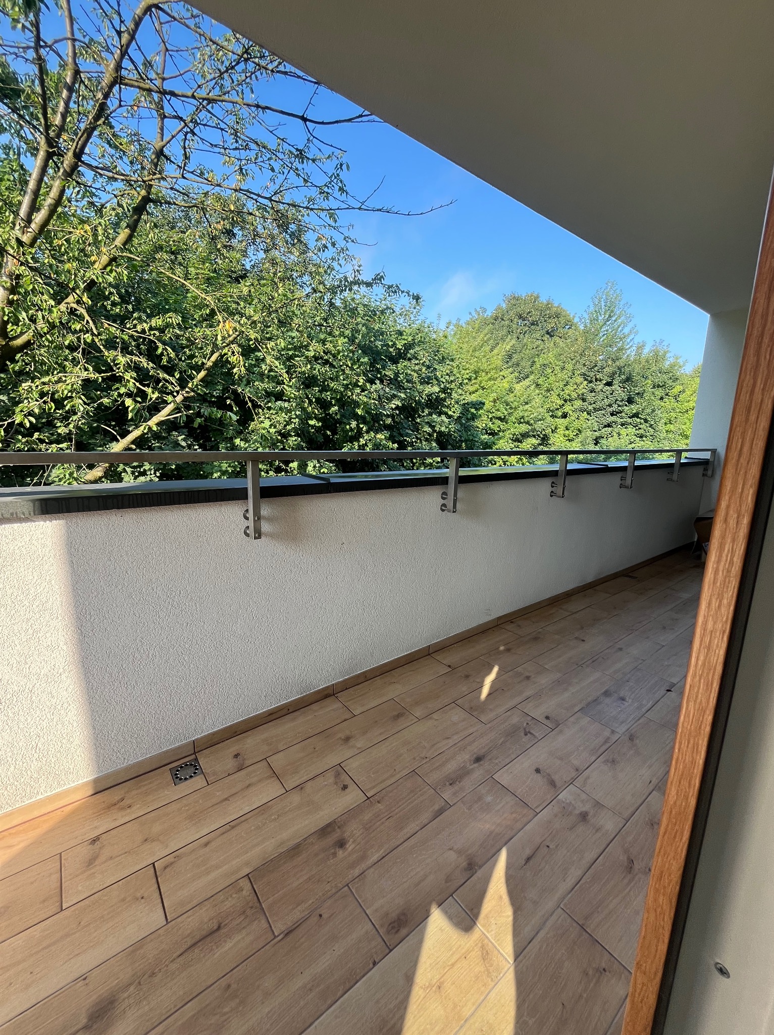 Wykończony balkon z drewnopodobnymi płytkami, metalową balustradą i widokiem na zieleń drzew.