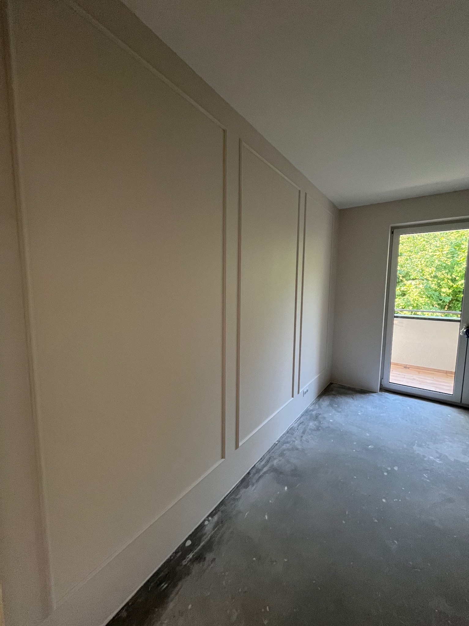 Ściana z dekoracyjnymi panelami w kolorze beżowym, surowa betonowa podłoga przygotowana pod wykończenie, widok na balkon z zielenią za oknem.