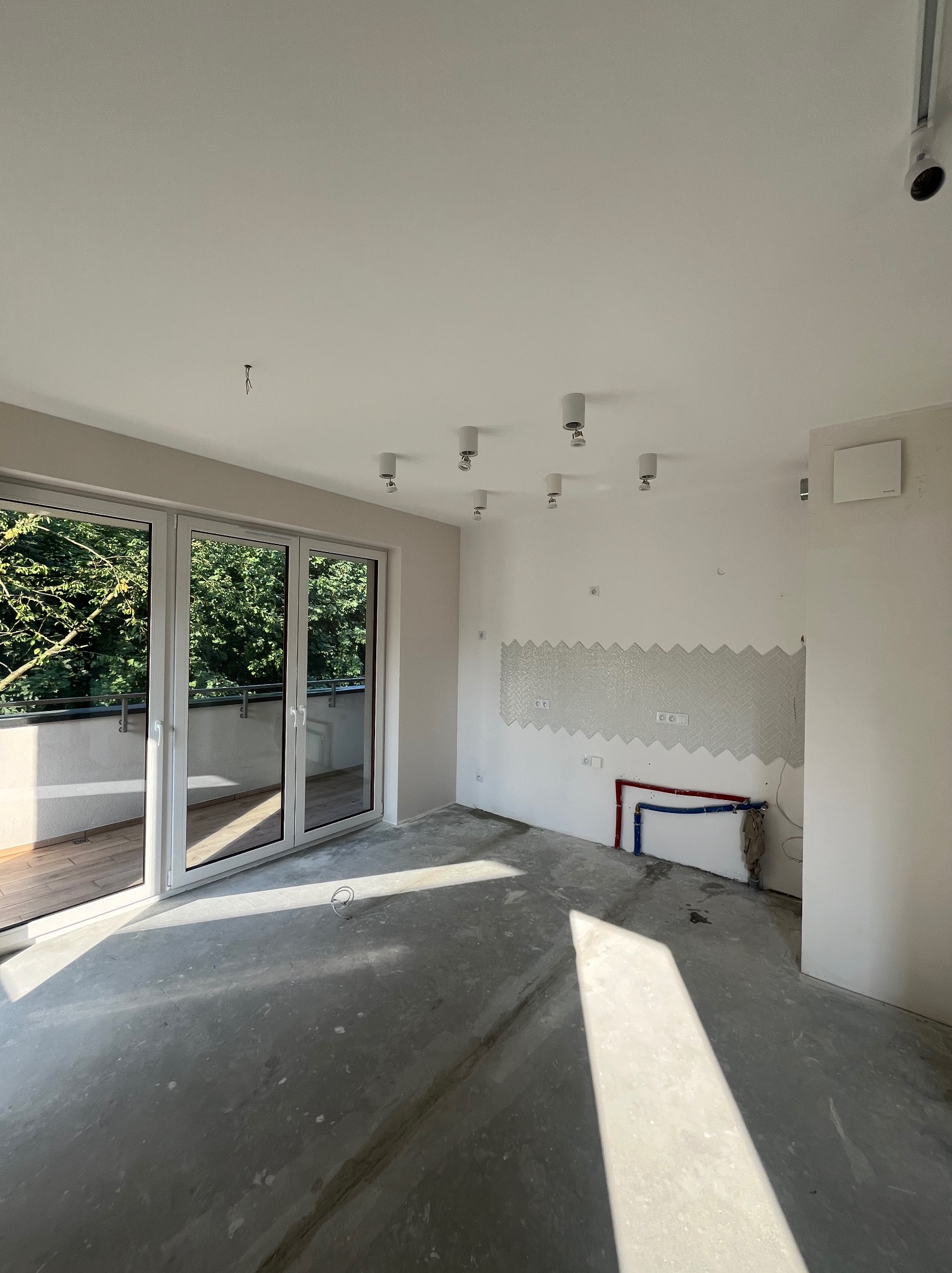 Wnętrze w trakcie prac: częściowo położona mozaika na ścianie, wyprowadzone instalacje hydrauliczne, surowa podłoga, widok na balkon z zielenią.