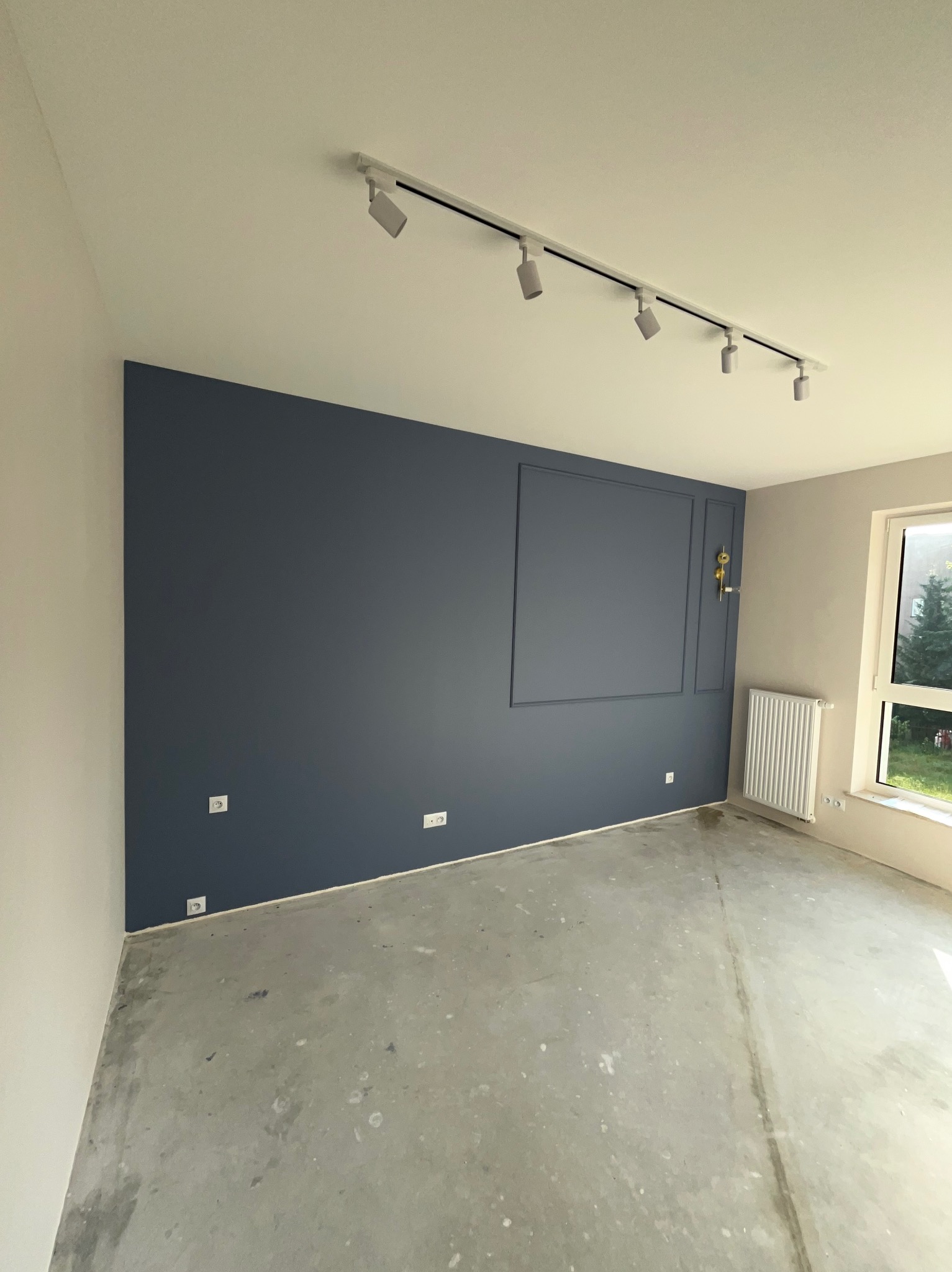Pokój po remoncie: granatowa ściana z dekoracyjną ramą, szara druga ściana, surowa betonowa podłoga, biały grzejnik i listwa oświetleniowa z reflektorami.