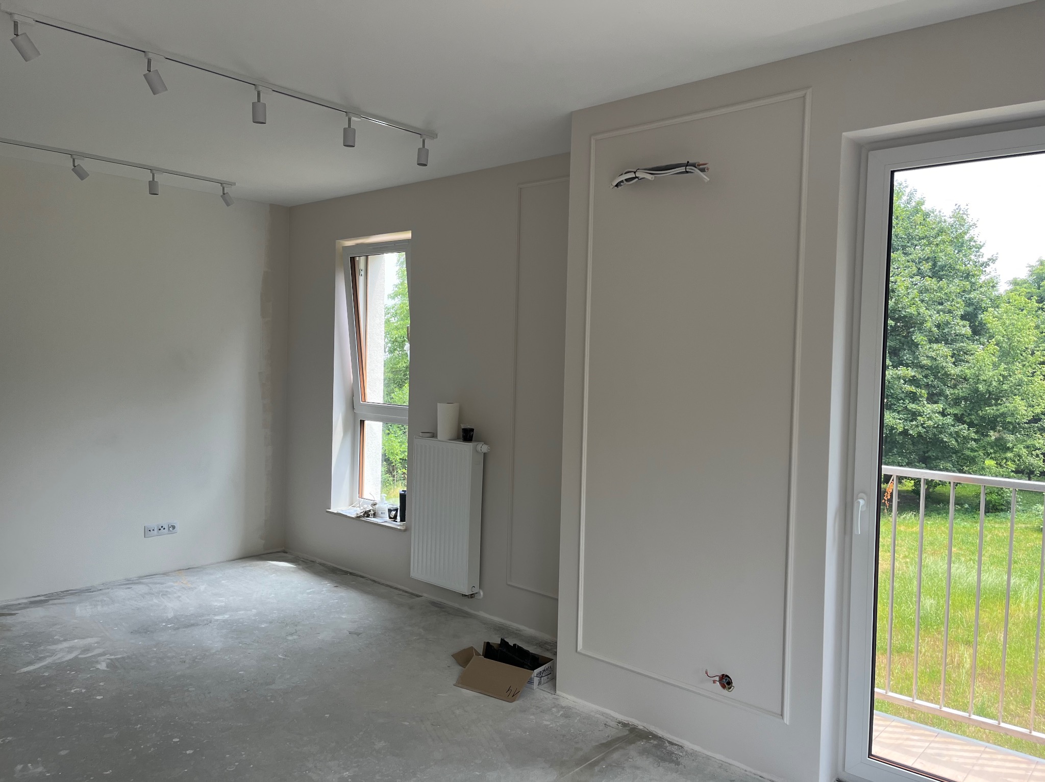 Pusty pokój w trakcie remontu z szarym betonowym podłożem, białymi ścianami z dekoracyjnymi listwami, grzejnikiem pod oknem i widocznymi kablami elektrycznymi wystającymi ze ściany.
