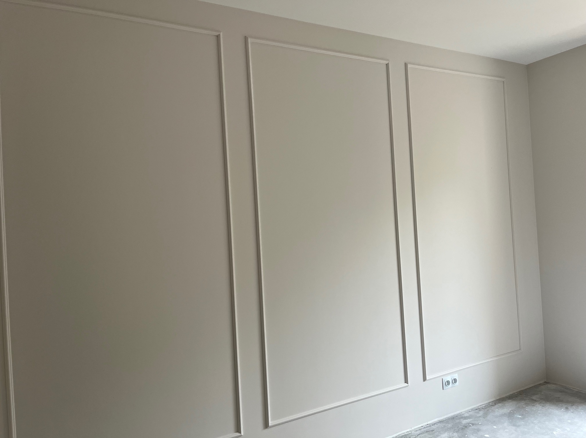 Ściana w jasnym odcieniu z dekoracyjnymi, pionowymi listwami tworzącymi panele. Widoczne gniazdka elektryczne na dole ściany.