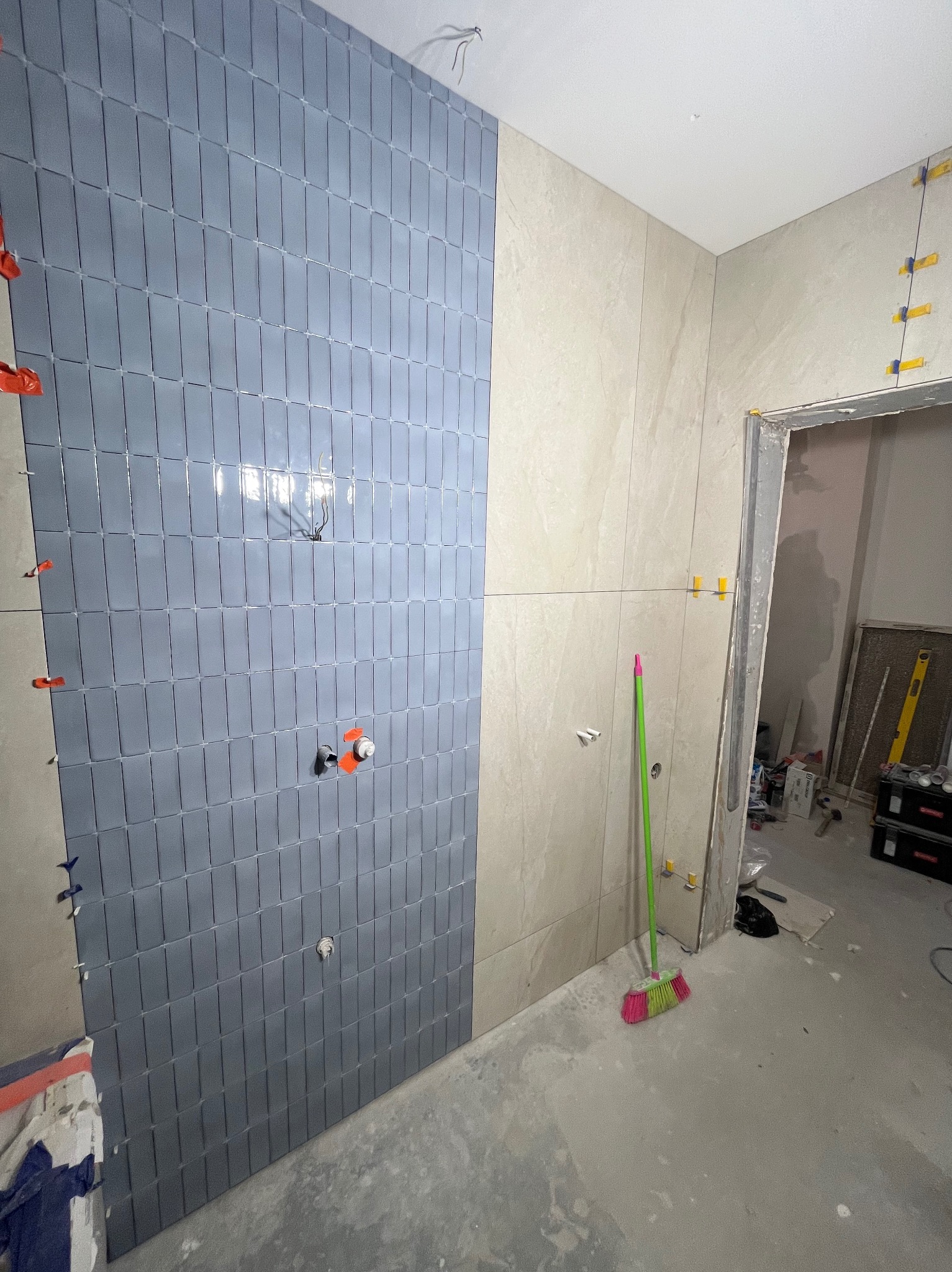 Ściana łazienki w trakcie układania płytek: szare, podłużne kafelki na jednej ścianie, beżowe płytki na drugiej, widoczne krzyżyki dystansowe i elementy instalacji wodnej, na podłodze leży miotła.