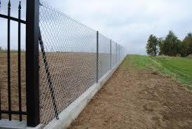 Długi, siatkowy płot na betonowej podmurówce, widok z boku, z czarnymi słupkami na początku, pole uprawne w tle.