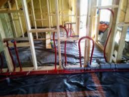 Czerwone rury instalacji grzewczej ułożone w pętle na czarnej folii, widoczne drewniane elementy konstrukcji szkieletowej budynku w trakcie budowy.