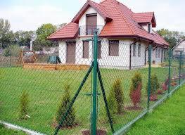 Zielone ogrodzenie z siatki na tle białego domu z czerwonym dachem, widoczne młode drzewka wzdłuż ogrodzenia i elementy placu zabaw w oddali.