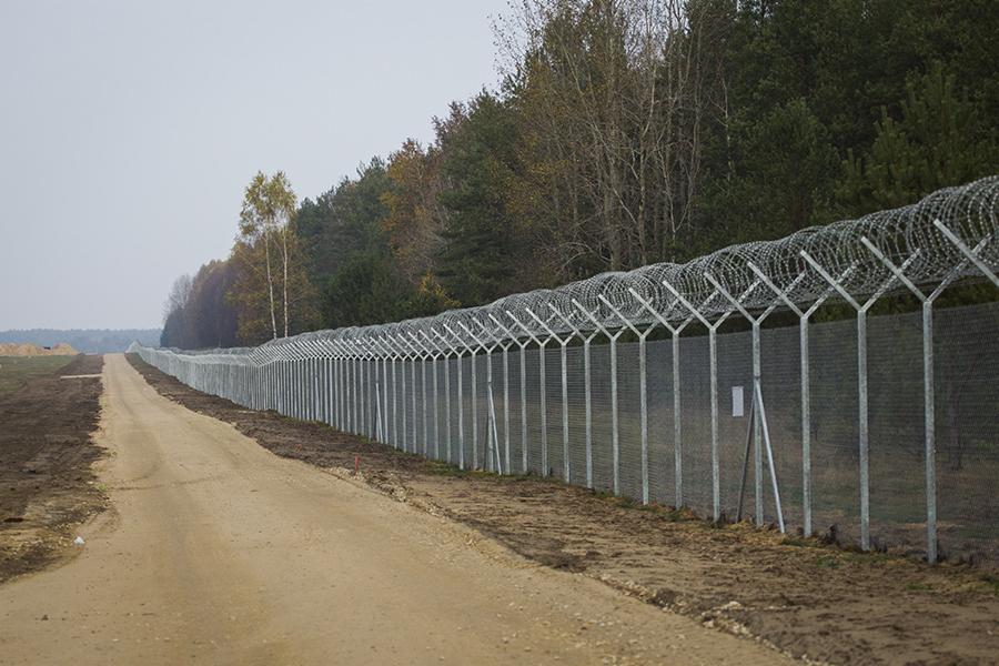 Długa metalowa siatka ogrodzeniowa z drutem kolczastym biegnąca wzdłuż polnej drogi, otoczona drzewami w tle.