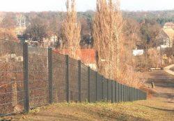 Długi, szary płot z siatki panelowej na nierównym terenie, widok z dołu, w tle drzewa bez liści i zabudowania.