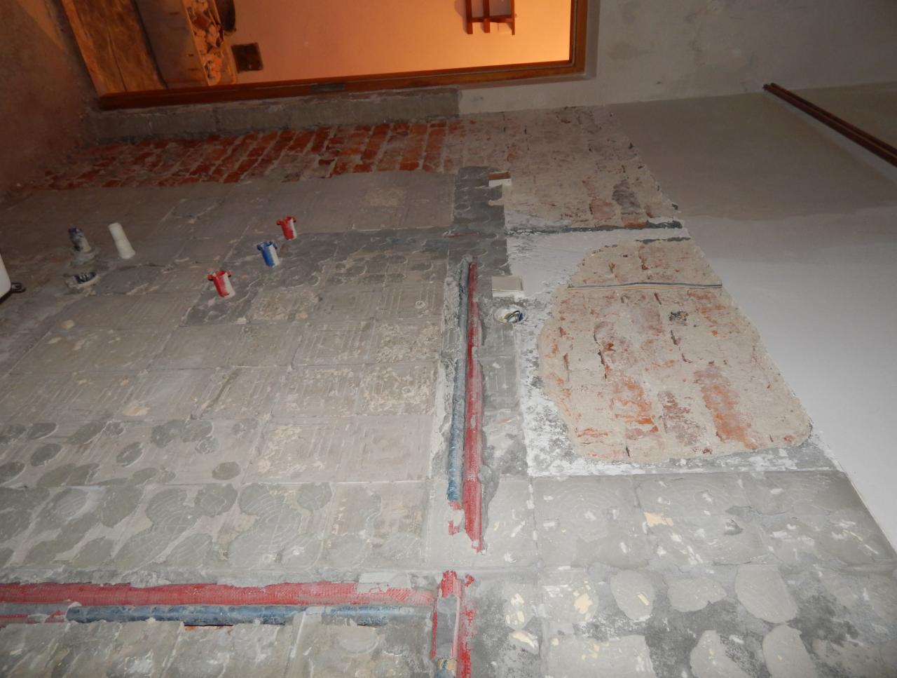 Fragment ściany z odsłoniętą czerwoną cegłą i częściowo usuniętym tynkiem, widoczne instalacje hydrauliczne w kolorze czerwonym i niebieskim, oraz elementy mocujące do rur.
