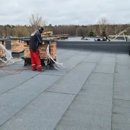 Pracownik w czerwonych spodniach i czarnej kurtce uszczelnia papą komin na płaskim dachu, butla gazowa obok, widok na las i domy w tle.