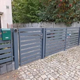 Nowoczesna, pozioma, szara brama wjazdowa z metalowymi przęsłami, skrzynka pocztowa w kolorze zielonym, kostka brukowa na podjeździe.