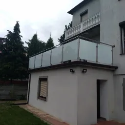 Nowoczesny balkon z przeszkloną konstrukcją, stalowe słupki, widok z zewnątrz na tle zieleni i domu z balustradą z białymi tralkami na wyższym poziomie.