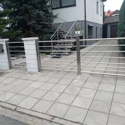 Balustrady Paweł Grzesiak - Nowoczesna, pozioma brama wjazdowa ze stali nierdzewnej z przesuwną furtką, zamontowana na betonowych słupkach przed domem z zewnętrznymi schodami również z balustradą ze stali nierdzewnej.