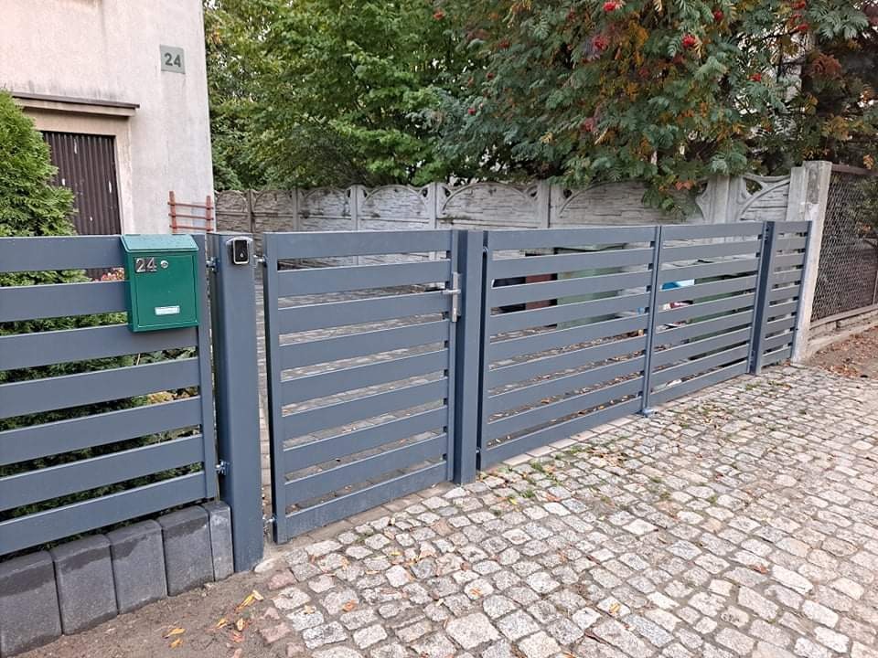 Nowoczesna, pozioma, szara brama wjazdowa z metalowymi przęsłami, skrzynka pocztowa w kolorze zielonym, kostka brukowa na podjeździe.