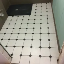 Układanie białych płytek z czarnym akcentem na podłodze w łazience, widoczny brodzik i żółte wiaderko z krzyżykami dystansowymi.