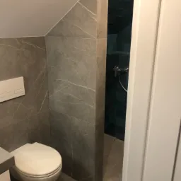 Nowoczesna łazienka z szarymi płytkami imitującymi marmur, podwieszana toaleta i kabina prysznicowa z ciemnozielonymi płytkami widoczna przez otwarte drzwi.