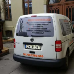 Biały samochód dostawczy z naklejką reklamową usług kominiarskich na tylnej szybie, zaparkowany przed budynkiem.