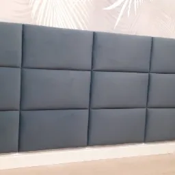 Tapicerowany panel ścienny w kolorze gołębim z wbudowanymi gniazdkami elektrycznymi, zamontowany na ścianie z delikatnym wzorem liści palmowych.